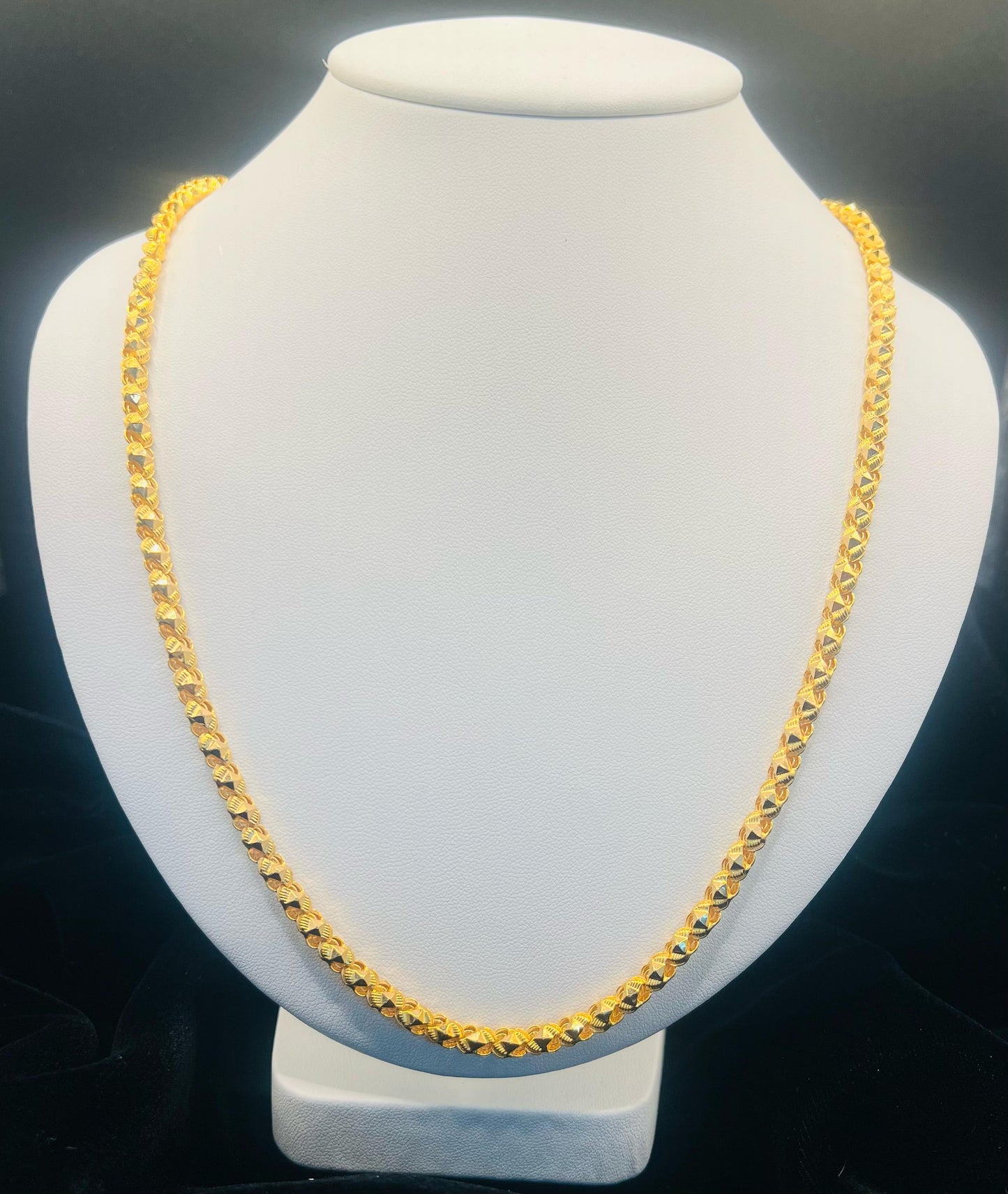A gold-toned chain necklace displayed on a white jewelry bust against a dark background.